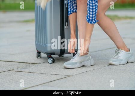 Primo piano delle mani delle donne che legano le scarpe in sneakers grigie accanto a una valigia grigia, spazio copia Foto Stock