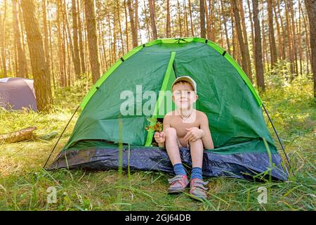 Il ragazzo siede nella tenda turistica nella foresta verde. Foto Stock