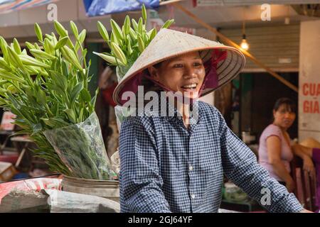 Donna sorridente che porta fiori in bicicletta nelle strade trafficate di Hanoi, Vietnam. Foto Stock