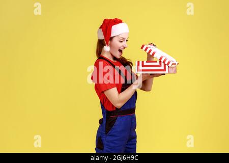 Vista laterale della donna Handy apre la scatola presente dal datore di lavoro, l'industria di servizio si congratula con i lavoratori con Natale, indossando tute e santa cappello. Indoo Foto Stock