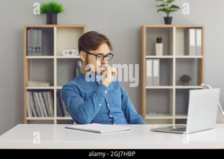 Ritratto di pensiero giovane concentrato mentre si siede in ufficio di fronte al portatile. Foto Stock