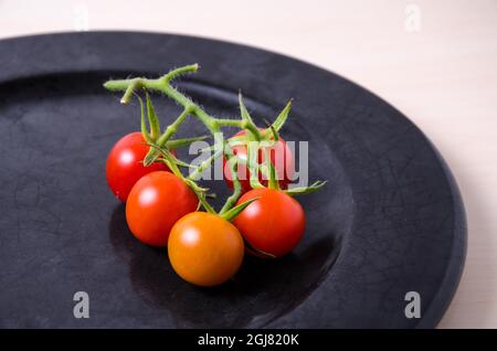 Pomodori ciliegini rossi maturi (Solanum lycopersicum var. Cerasiforme) con gambo verde su un piatto scuro. Vista dall'alto. Foto Stock