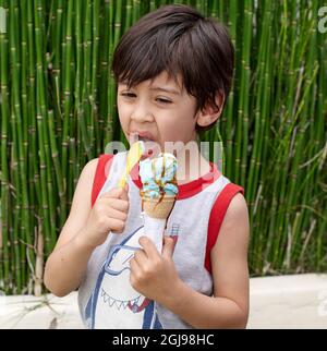 Ragazzino di brunette che lecca il cono gelato e succhia un cucchiaio, indossando una camicia grigia senza maniche in estate. Piante verdi su sfondo. Verticale Foto Stock