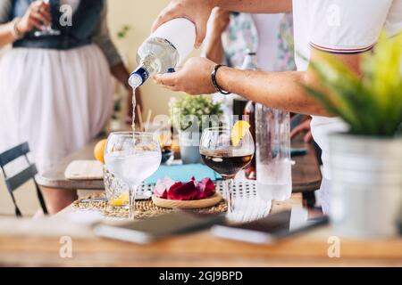 Mani di uomo che versano alcool in vetro preparare un cocktail o una bevanda rinfrescante sul tavolo da pranzo mentre la donna beve in background. Preparazione d Foto Stock