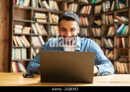 Sorpreso positivo sorridente ragazzo di freelance indiano utilizzando il portatile seduta in ufficio o biblioteca, stupito multirazziale est maschio dipendente ha ricevuto un buon affare o notifica Foto Stock