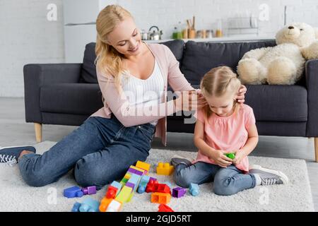 donna sorridente che regola i capelli della figlia giocando con i blocchi di costruzione sul pavimento a casa Foto Stock