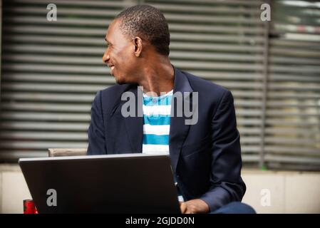 bell'uomo seduto su una panchina vicino all'azienda utilizzando un computer portatile e guardando lateralmente sorridente Foto Stock