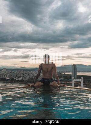 La vista dalla piscina sul tetto dell'hotel e l'uomo seduto sul bordo della piscina sul tetto. Vista pittoresca, bellissimo cielo Foto Stock