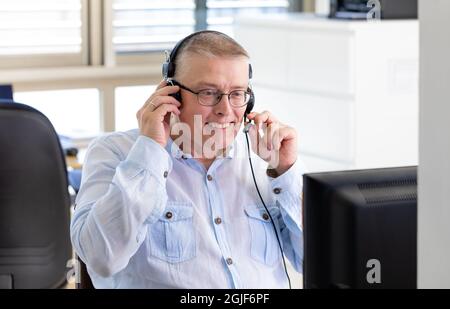 Un uomo lavora come manager in un ufficio e parla con i clienti su Internet attraverso un computer con cuffie e microfono. Uomo di aspetto europeo in una maglietta blu si siede in ufficio con la schiena alla finestra. Foto Stock