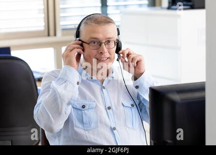 Un uomo lavora come manager in un ufficio e parla con i clienti su Internet attraverso un computer con cuffie e microfono. Uomo di aspetto europeo in una maglietta blu si siede in ufficio con la schiena alla finestra. Foto Stock