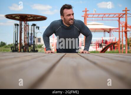 Un bell'uomo di mezza età si spinge dal pavimento sul terreno sportivo sullo sfondo dei simulatori, guarda via sull'espirazione, gode di mornina Foto Stock