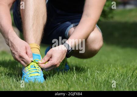 Uomo seduto su erba e allacciatura merletti su scarpe da ginnastica blu primo piano Foto Stock