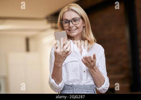 Chiamata online. Bella donna anziana in casual indossare sorridente e parlare mentre si fa una videochiamata utilizzando il suo smartphone, in piedi a casa Foto Stock