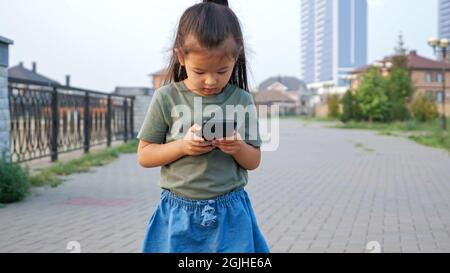 Piccola ragazza asiatica digita messaggio sul moderno cellulare in piedi all'aperto Foto Stock