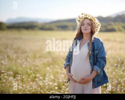 Bella donna incinta in un abito, giacca in denim e una corona di margherite rilassante fuori nel parco. Gravidanza sana e concetto di viaggio. Foto Stock