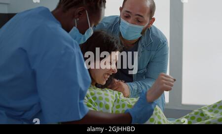 Giovane donna in travaglio che partorisce il bambino nel reparto ospedaliero con il sostegno del marito e dell'infermiere di ostetricia afroamericana. Paziente in gravidanza con contrazioni che spingono per il parto Foto Stock