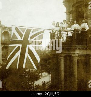 Il 22 giugno 1897, il giorno del Giubileo dei Diamanti della Regina Vittoria, giovani donne britanniche vestite in modo intelligente, alunni di una scuola di finitura in stile svizzero a Wiesbaden, Germania, celebrano il 60° anniversario della sua adesione al trono del Regno Unito, volando una grande bandiera dell'Unione o Union Jack da un balcone. Foto Stock