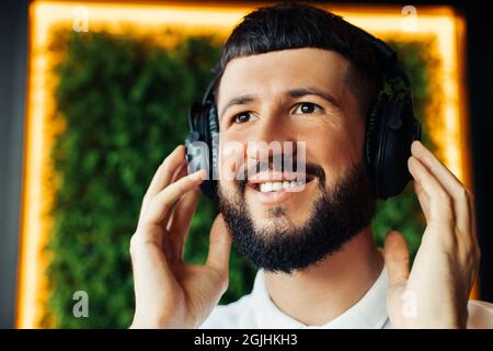 Sorridente giovane con barba che ascolta musica con le cuffie, giovane uomo in abiti casual che riposa e ascolta musica in un bar Foto Stock