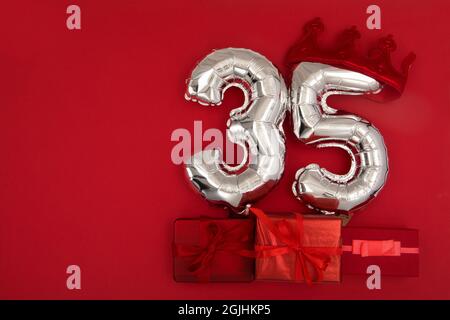 Palloncini d'aria in lamina d'argento che mostrano il numero di trentacinque 35 posti su sfondo rosso con i regali avvolti per il concetto di anniversario di compleanno Foto Stock