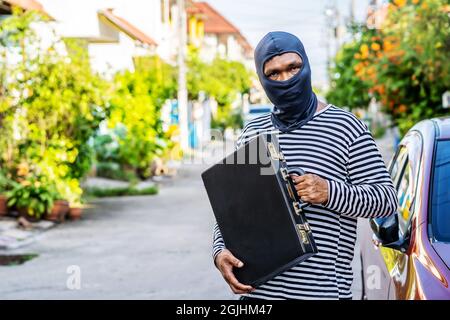 Il ladro maschio indossa una maschera nera, sorridendo felicemente dopo aver rubato la valigetta con i soldi che vuole Foto Stock