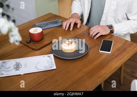 vista dall'alto dell'uomo vicino alla torta al limone, al cappuccino, al menu e allo smartphone con schermo vuoto sul tavolo Foto Stock