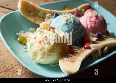 Primo piano di dolce dolce banana split dessert ad angolo alto con palline colorate di gelato decorate con spolverini e dragee servito su piatto su tavola di legno Foto Stock