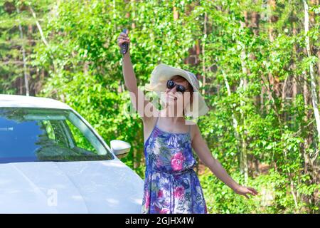 Bella gioiosa donna di fronte a un'auto bianca con una chiave in mano in una giornata estiva di sole sullo sfondo di alberi verdi. Messa a fuoco selettiva Foto Stock