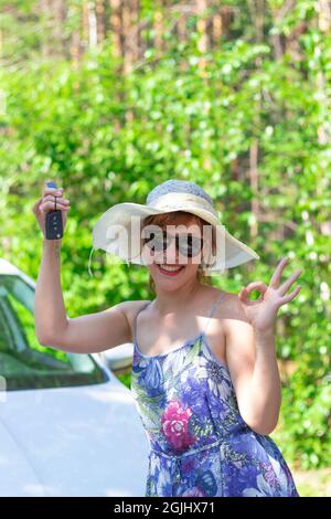 Bella gioiosa donna di fronte a un'auto bianca con una chiave in mano in una giornata estiva di sole sullo sfondo di alberi verdi. Messa a fuoco selettiva Foto Stock