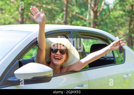 Una bella gioiosa donna guarda fuori dalla finestra dell'auto con una chiave in mano in una giornata estiva soleggiata sullo sfondo di alberi verdi. Messa a fuoco selettiva Foto Stock