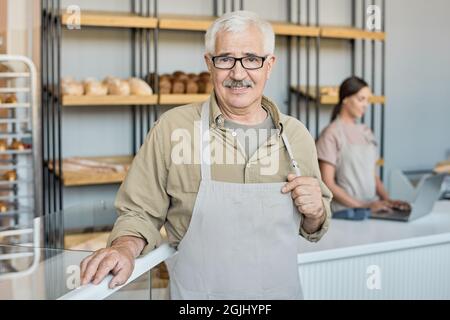 Ritratto di buon panettiere invecchiato con baffi che tiene la trappola di grembiule mentre si posa contro esposizione con assortimento di pane fresco Foto Stock