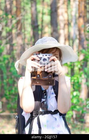 Giovane ragazza turistica in un cappello di paglia con uno zaino e una macchina fotografica nella foresta sullo sfondo di alberi verdi in una giornata estiva di sole. Selettivo fo Foto Stock