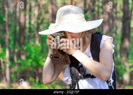 Giovane ragazza turistica in un cappello di paglia con uno zaino e una macchina fotografica nella foresta sullo sfondo di alberi verdi in una giornata estiva di sole. Selettivo fo Foto Stock