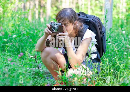 Giovane ragazza turistica con uno zaino e una macchina fotografica nella foresta su uno sfondo di erba verde in una giornata estiva di sole. Messa a fuoco selettiva Foto Stock