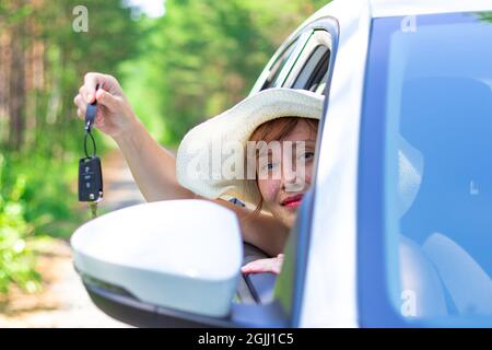 Una bella gioiosa donna guarda fuori dalla finestra dell'auto con una chiave in mano in una giornata estiva soleggiata sullo sfondo di alberi verdi. Messa a fuoco selettiva Foto Stock