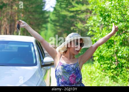 Bella gioiosa donna di fronte a un'auto bianca con una chiave in mano in una giornata estiva di sole sullo sfondo di alberi verdi. Messa a fuoco selettiva Foto Stock