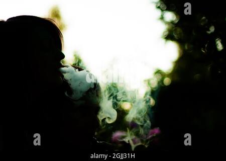 Silhouette di una persona che fuma hookah e che soffia il fumo su sfondo sfocato scuro e chiaro Foto Stock