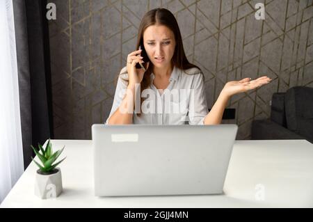 Donna irritata che parla al telefono mentre si siede alla scrivania in ufficio a casa e si utilizza un computer portatile. Turbato e pensivo freelance femmina parlare sullo smartphone, non capire qualcosa correttamente Foto Stock