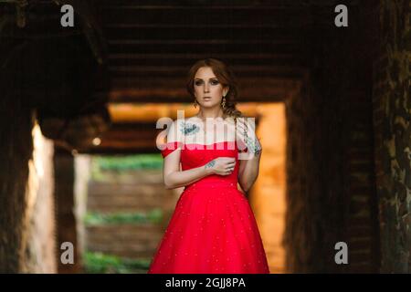 Attraente donna tatuata rossa in lungo vestito rosso e diadema su sfondo sfocato castello medievale Foto Stock