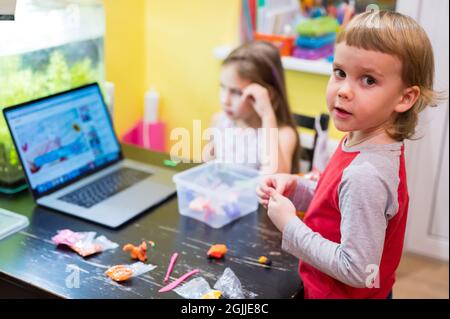 bambini piccoli, ragazza e ragazzo, impegnati nella modellazione creativa da argilla o plastilina in una stanza a un tavolo, guardando una lezione online di classe su un computer o. Foto Stock