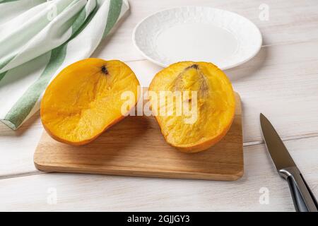 Mango molto maturo tagliato a metà su un tagliere, coltello da frutta e piatto su tavola di legno bianco. Mango giallo succoso delizioso. Frutta tropicale dolce. Foto Stock