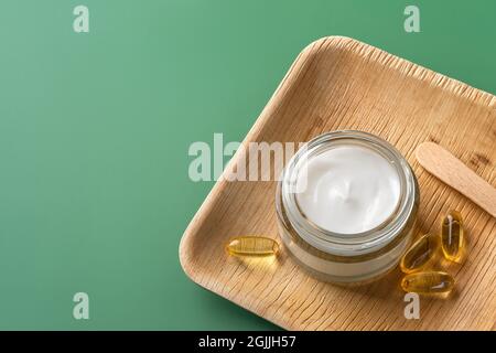 Crema per la cura della pelle di bellezza e capsule di olio essenziali su un vassoio di legno su sfondo verde pastello. Crema idratante bianca in un vaso di vetro. Spazio di copia. Foto Stock