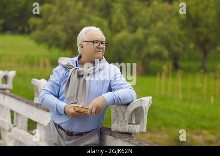 L'uomo anziano felice con un libro nelle sue mani sta riposando nel parco all'aperto. Foto Stock