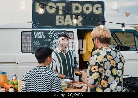 Giovani che cucinano e vendono hot dog ai giovani mentre si levano in piedi all'aperto Foto Stock
