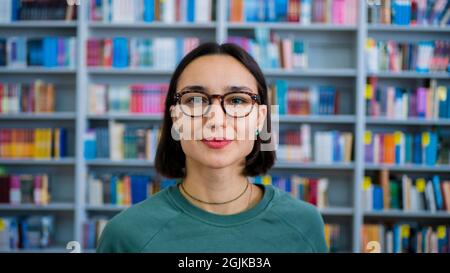 Primo piano ritratto di una giovane studentessa sullo sfondo di librerie nella biblioteca universitaria. Giovane donna millenaria che guarda Foto Stock