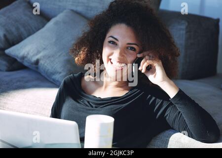 Felice giovane donna che chiacchiera sul suo smartphone mentre si rilassa sul pavimento al sole a casa a distanza lavorando su un computer portatile Foto Stock