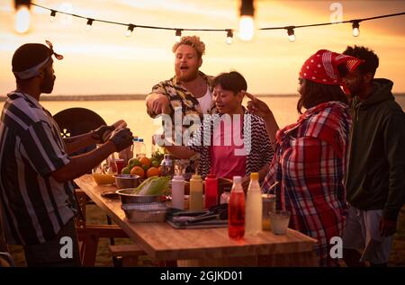 Giovani che mangiano fast food al tavolo durante una festa in spiaggia all'aperto Foto Stock