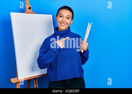 Bella donna ispanica con i capelli corti che tengono le spazzole vicino al cavalletto sorridendo felice puntando con la mano e il dito Foto Stock