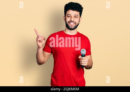 Giovane uomo arabo con la barba cantare canzone con microfono sorridente felice puntando con mano e dito a lato Foto Stock