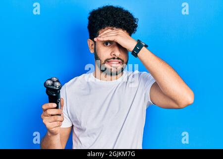 Giovane uomo arabo con barba che tiene la macchina elettrica del rasoio stressato e frustrato con mano sulla testa, sorpreso e faccia arrabbiata Foto Stock