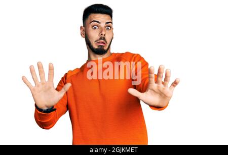 Giovane ispanico con barba con maglione arancione casual paura e terrorizzato con paura espressione stop gesto con le mani, gridando in shock. pani Foto Stock